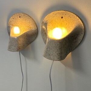 May include: A pair of wall-mounted ceramic sconces with a speckled cream and brown finish. Each lamp features a curved, shell-like shape with a central opening that houses a glowing light bulb. A thin white power cord hangs from each fixture.