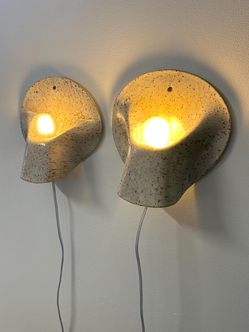 May include: A pair of wall-mounted ceramic sconces with a speckled cream and brown finish. Each lamp features a curved, shell-like shape with a central opening that houses a glowing light bulb. A thin white power cord hangs from each fixture.