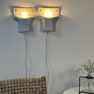 May include: A pair of wall-mounted sconces featuring a smooth, light gray ceramic finish with a flared, architectural shape. Each fixture emits a warm, glowing light from a central bulb, with a thin white power cord extending downward from each unit.