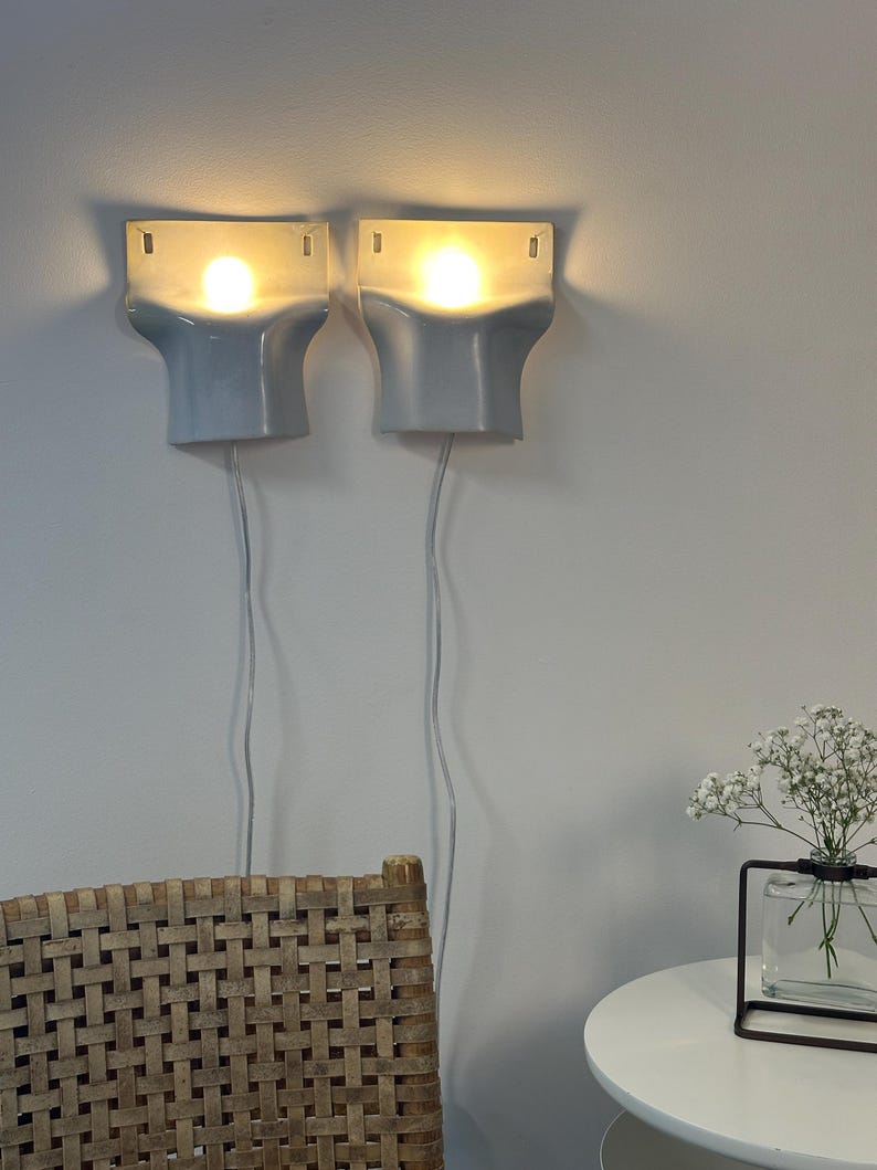 May include: A pair of wall-mounted sconces featuring a smooth, light gray ceramic finish with a flared, architectural shape. Each fixture emits a warm, glowing light from a central bulb, with a thin white power cord extending downward from each unit.