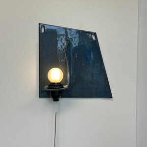 May include: A modern wall sconce featuring a blue, geometric metal backplate with a curved metal arm. A round, illuminated light bulb is set in a black socket. A silver cord extends downward from the fixture.