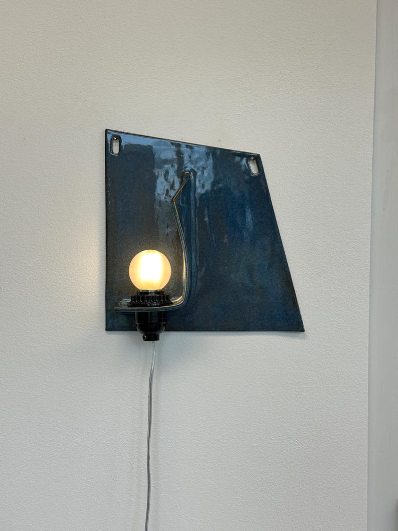 May include: A modern wall sconce featuring a blue, geometric metal backplate with a curved metal arm. A round, illuminated light bulb is set in a black socket. A silver cord extends downward from the fixture.