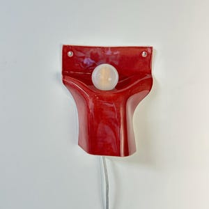 Pode incluir: Uma lumin&aacute;ria de parede com uma base de cer&acirc;mica vermelha brilhante de formato escultural e alargado. O acess&oacute;rio inclui uma l&acirc;mpada branca redonda centrada na parte superior e dois pequenos orif&iacute;cios para montagem. Um fio fino claro pende.