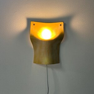May include: A wall-mounted sconce featuring a curved, golden-yellow ceramic shade with a concave center. A bright, warm light bulb is positioned in the middle, casting a soft glow against the wall. A thin power cord hangs from the bottom.