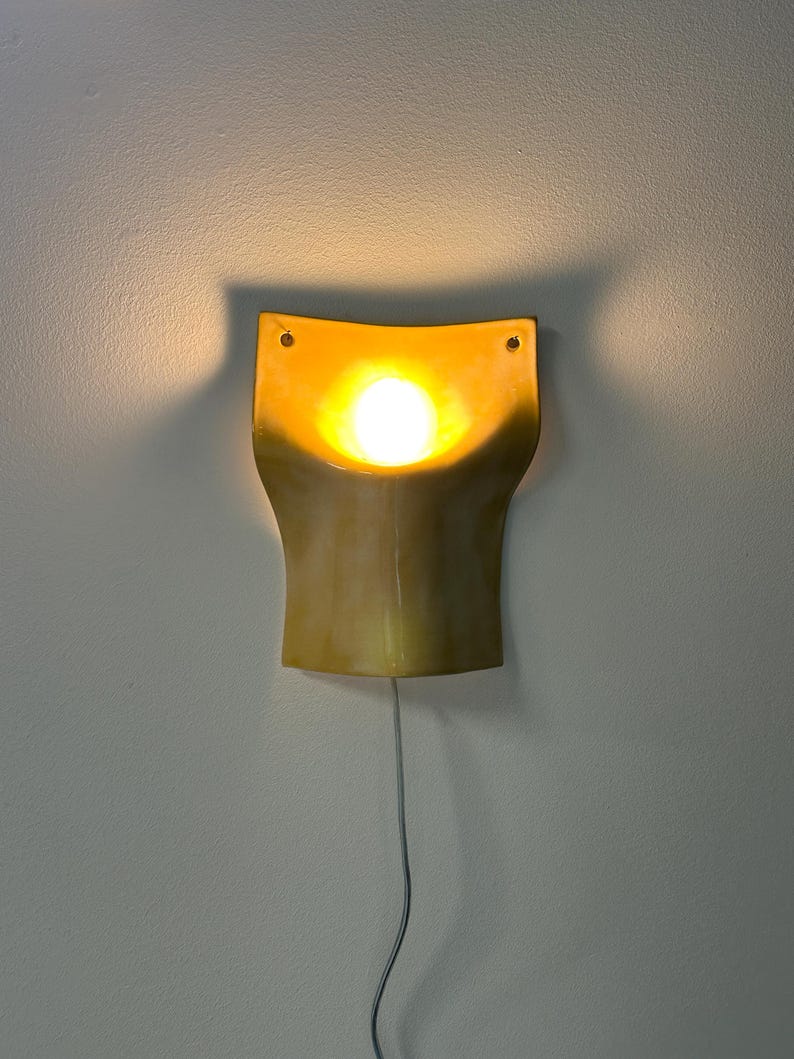 May include: A wall-mounted sconce featuring a curved, golden-yellow ceramic shade with a concave center. A bright, warm light bulb is positioned in the middle, casting a soft glow against the wall. A thin power cord hangs from the bottom.