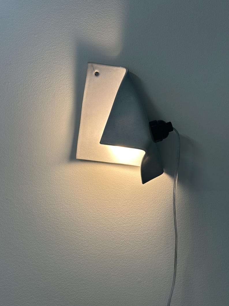May include: A white ceramic wall sconce with a folded, paper-like design that emits a warm, downward-facing light. The fixture features a black socket attached to the side, connected to a thin, transparent electrical cord hanging downward.