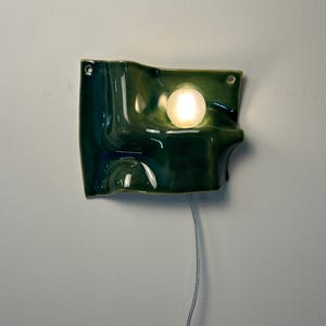 May include: A wall-mounted lamp featuring a glossy, dark green ceramic shade with an undulating, wave-like surface. The fixture includes a single, round, frosted white light bulb emitting a warm glow and a thin, light-colored electrical cord.
