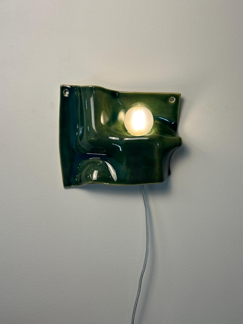 May include: A wall-mounted lamp featuring a glossy, dark green ceramic shade with an undulating, wave-like surface. The fixture includes a single, round, frosted white light bulb emitting a warm glow and a thin, light-colored electrical cord.