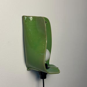 Pu&ograve; includere: Una lampada da parete con un paralume in ceramica verde brillante dalla forma organica e curva. L'apparecchio include una lampadina sferica bianca, un portalampada nero e un cavo di alimentazione nero che scende verso il basso dalla base.