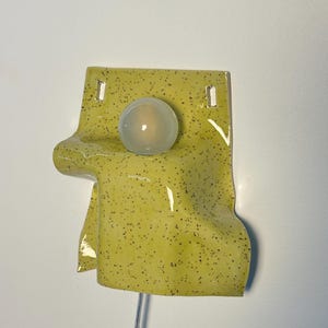 Puede incluir: Un aplique de pared con una pantalla de cer&aacute;mica brillante de color chartreuse con un patr&oacute;n de motas oscuras. La pantalla tiene una apariencia drapeada y plegada con dos orificios de montaje rectangulares en la parte superior y una bombilla redonda esmerilada.