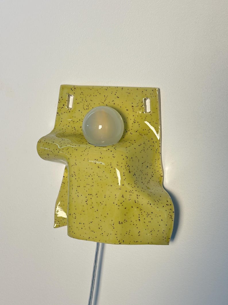 Puede incluir: Un aplique de pared con una pantalla de cer&aacute;mica brillante de color chartreuse con un patr&oacute;n de motas oscuras. La pantalla tiene una apariencia drapeada y plegada con dos orificios de montaje rectangulares en la parte superior y una bombilla redonda esmerilada.