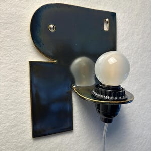 May include: A wall-mounted lamp featuring a dark blue, glossy ceramic backplate with a metallic gold edge. A black socket extends from the centre, holding a small, round, frosted white light bulb. A thin, clear power cord hangs from the bottom.