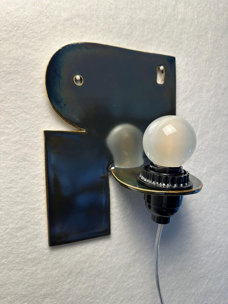 May include: A wall-mounted lamp featuring a dark blue, glossy ceramic backplate with a metallic gold edge. A black socket extends from the centre, holding a small, round, frosted white light bulb. A thin, clear power cord hangs from the bottom.