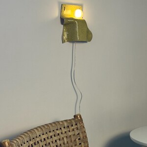 Puede incluir: Un aplique de pared con una pantalla de cer&aacute;mica de color verde claro moteado y forma curva y org&aacute;nica. Una bombilla redonda e iluminada se sit&uacute;a en la parte superior de la pieza de cer&aacute;mica, conectada por un cable el&eacute;ctrico fino y transparente.