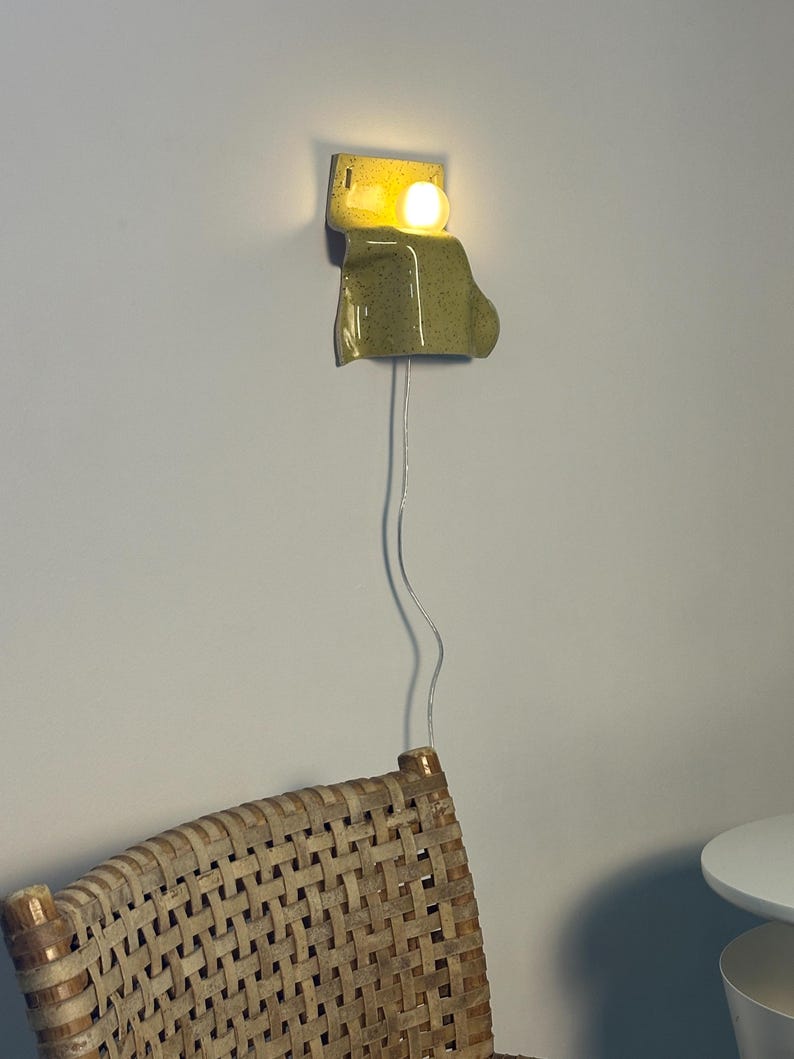 Puede incluir: Un aplique de pared con una pantalla de cer&aacute;mica de color verde claro moteado y forma curva y org&aacute;nica. Una bombilla redonda e iluminada se sit&uacute;a en la parte superior de la pieza de cer&aacute;mica, conectada por un cable el&eacute;ctrico fino y transparente.