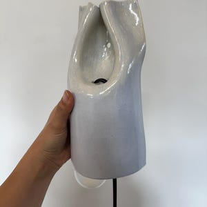 May include: A wall-mounted ceramic light fixture with a glossy, pale lavender-grey finish. The object features an organic, folded, and sculptural form with a central opening that reveals a black electrical cord and a small circular light source.