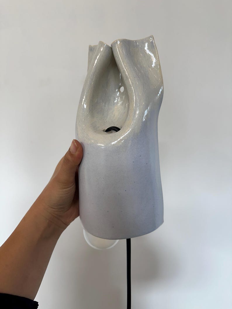 May include: A wall-mounted ceramic light fixture with a glossy, pale lavender-grey finish. The object features an organic, folded, and sculptural form with a central opening that reveals a black electrical cord and a small circular light source.