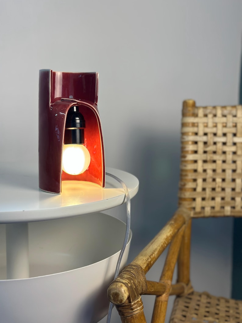 May include: A deep red ceramic table lamp with a curved, cylindrical body and an open archway design. The lamp features a black socket holding a glowing, round white light bulb and includes a clear power cord extending from the base.