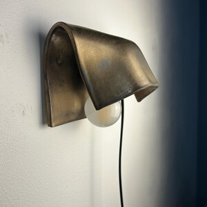 Pode incluir: Um candeeiro de parede com um abajur curvo em metal bronzeado com acabamento envelhecido. Uma l&acirc;mpada esf&eacute;rica branca est&aacute; posicionada sob o abajur e um fio de alimenta&ccedil;&atilde;o preto fino desce a partir da lumin&aacute;ria.
