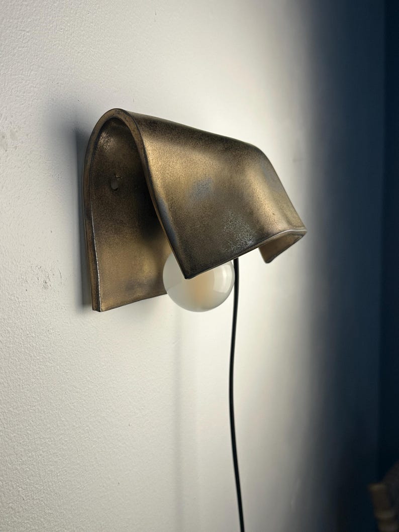 Pode incluir: Um candeeiro de parede com um abajur curvo em metal bronzeado com acabamento envelhecido. Uma l&acirc;mpada esf&eacute;rica branca est&aacute; posicionada sob o abajur e um fio de alimenta&ccedil;&atilde;o preto fino desce a partir da lumin&aacute;ria.