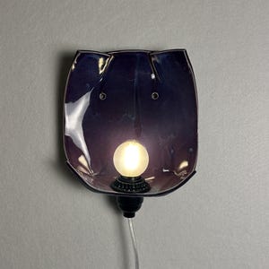 May include: A wall-mounted lamp featuring a deep purple, glossy ceramic shade with a curved, organic shape. The fixture includes a central, glowing frosted light bulb set into a black socket, with a clear power cord extending downwards from the base.