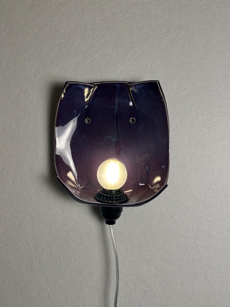 May include: A wall-mounted lamp featuring a deep purple, glossy ceramic shade with a curved, organic shape. The fixture includes a central, glowing frosted light bulb set into a black socket, with a clear power cord extending downwards from the base.