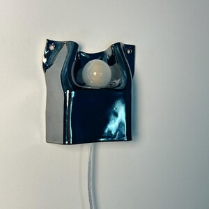 May include: A wall-mounted lamp featuring a dark, reflective, metallic-finished ceramic base with an irregular, fluid shape. A single, round, frosted white light bulb is centered in the upper cutout, with a clear power cord extending downward.