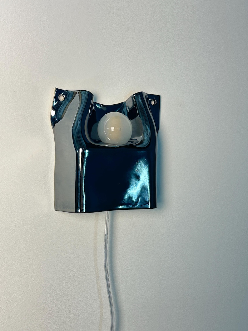 May include: A wall-mounted lamp featuring a dark, reflective, metallic-finished ceramic base with an irregular, fluid shape. A single, round, frosted white light bulb is centered in the upper cutout, with a clear power cord extending downward.
