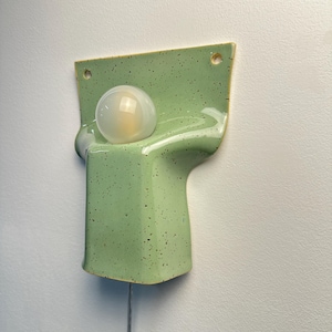 May include: A wall-mounted light fitting featuring a glossy, light green ceramic base with dark speckles. The base has a curved, moulded shape with two circular holes at the top for mounting and a single round, frosted glass light bulb centred.