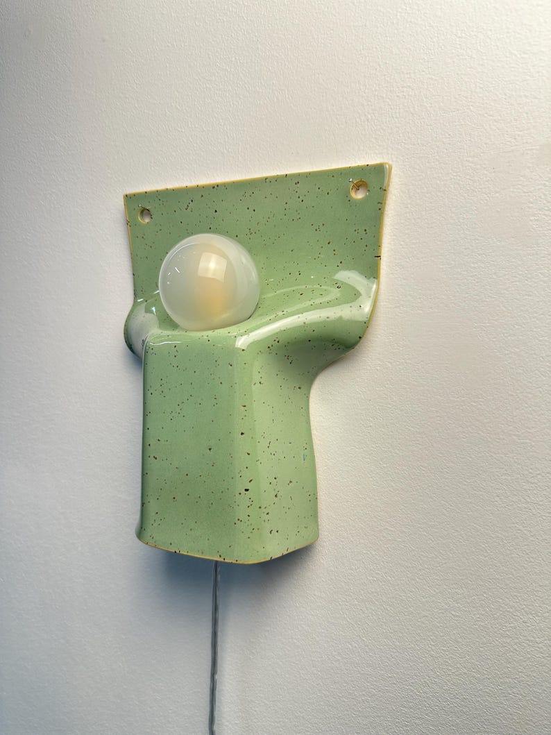 May include: A wall-mounted light fitting featuring a glossy, light green ceramic base with dark speckles. The base has a curved, moulded shape with two circular holes at the top for mounting and a single round, frosted glass light bulb centred.