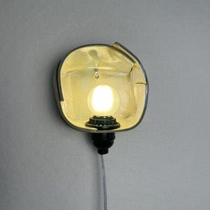 Puede incluir: Un aplique de pared con una pantalla reflectante met&aacute;lica redondeada en tono dorado con un borde irregular y plegado. Una bombilla esf&eacute;rica brillante est&aacute; centrada dentro de la pantalla, unida a un casquillo negro y un cable fino.
