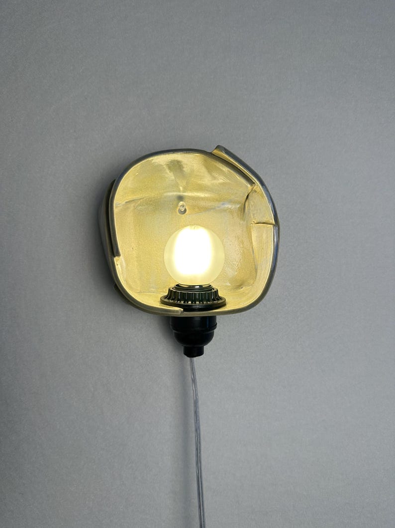 Puede incluir: Un aplique de pared con una pantalla reflectante met&aacute;lica redondeada en tono dorado con un borde irregular y plegado. Una bombilla esf&eacute;rica brillante est&aacute; centrada dentro de la pantalla, unida a un casquillo negro y un cable fino.
