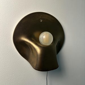 May include: A wall-mounted lamp featuring a dark bronze-toned, organic, curved metal shade with a smooth, matte finish. A single, round, frosted white glass light bulb is positioned in the center of the shade. A thin, light-colored power cord extends downward.