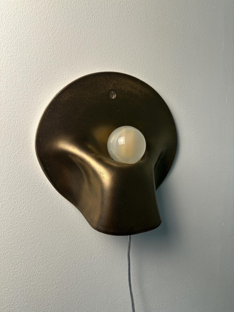 May include: A wall-mounted lamp featuring a dark bronze-toned, organic, curved metal shade with a smooth, matte finish. A single, round, frosted white glass light bulb is positioned in the center of the shade. A thin, light-colored power cord extends downward.