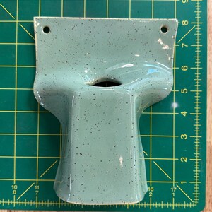 May include: A light green ceramic wall planter with dark speckles, featuring a wide top section with two mounting holes and a central opening. The object is positioned on a green cutting mat with a grid, showing a width of approximately 15 cm.