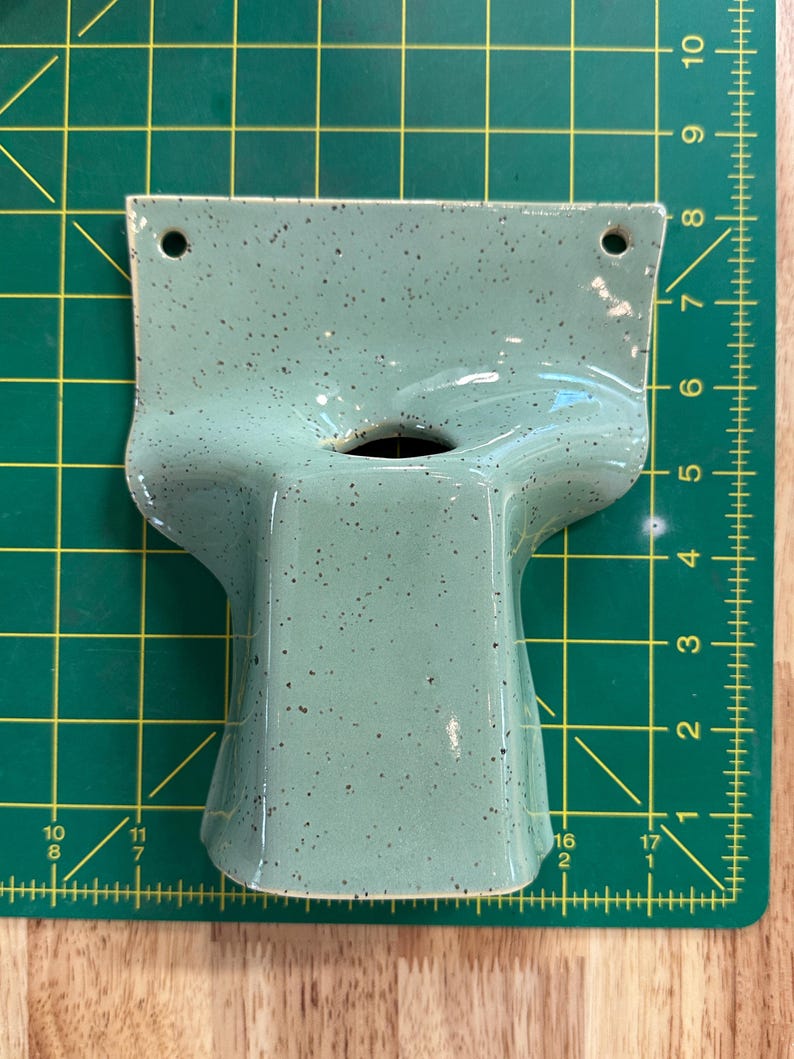 May include: A light green ceramic wall planter with dark speckles, featuring a wide top section with two mounting holes and a central opening. The object is positioned on a green cutting mat with a grid, showing a width of approximately 15 cm.