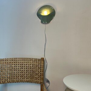 May include: A sage green wall-mounted lamp with a curved, shell-like shade and a warm glowing light bulb. The lamp features a thin white power cord that hangs vertically down the wall, equipped with a small inline switch for operation.
