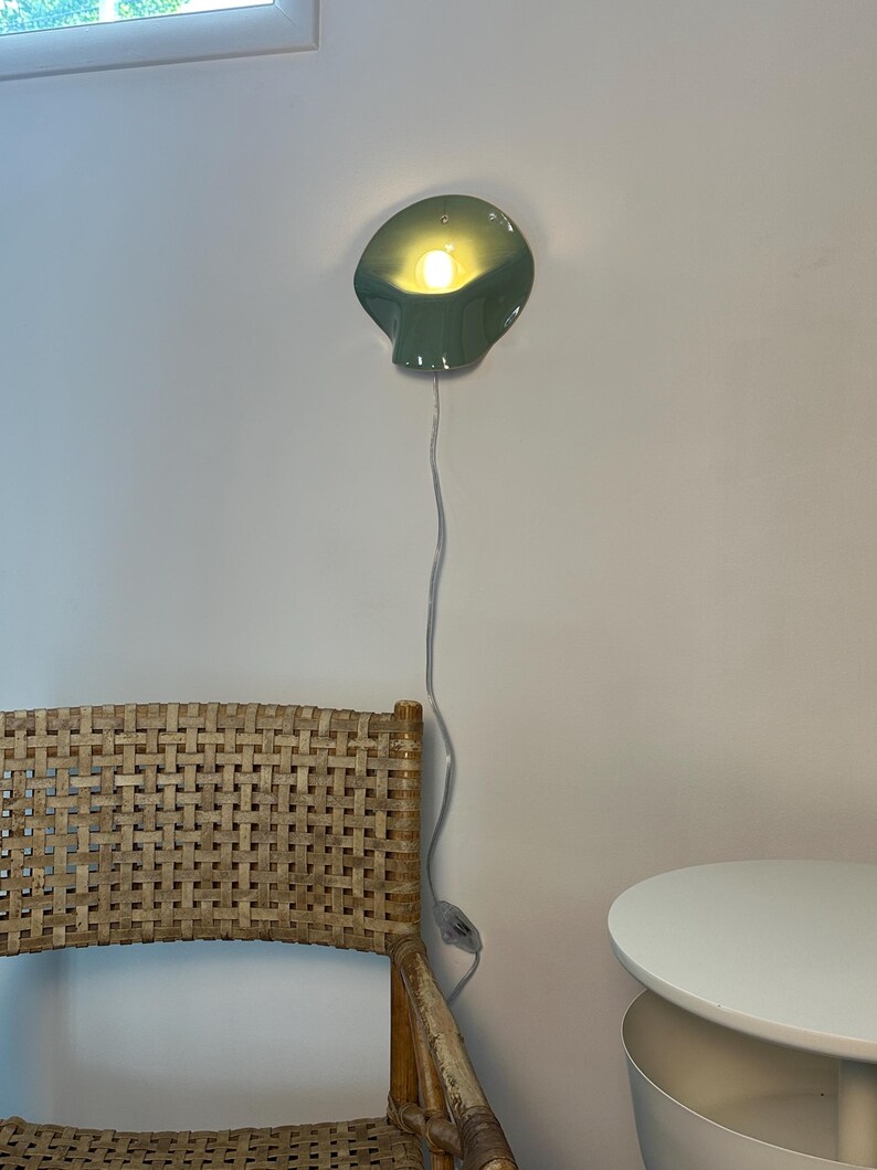 May include: A sage green wall-mounted lamp with a curved, shell-like shade and a warm glowing light bulb. The lamp features a thin white power cord that hangs vertically down the wall, equipped with a small inline switch for operation.