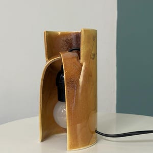 Puede incluir: Una l&aacute;mpara de mesa cil&iacute;ndrica de cer&aacute;mica con un esmalte amarillo mostaza brillante. La estructura presenta una abertura vertical que revela un casquillo negro y una bombilla redonda blanca. Un cable de alimentaci&oacute;n negro sale de la base.