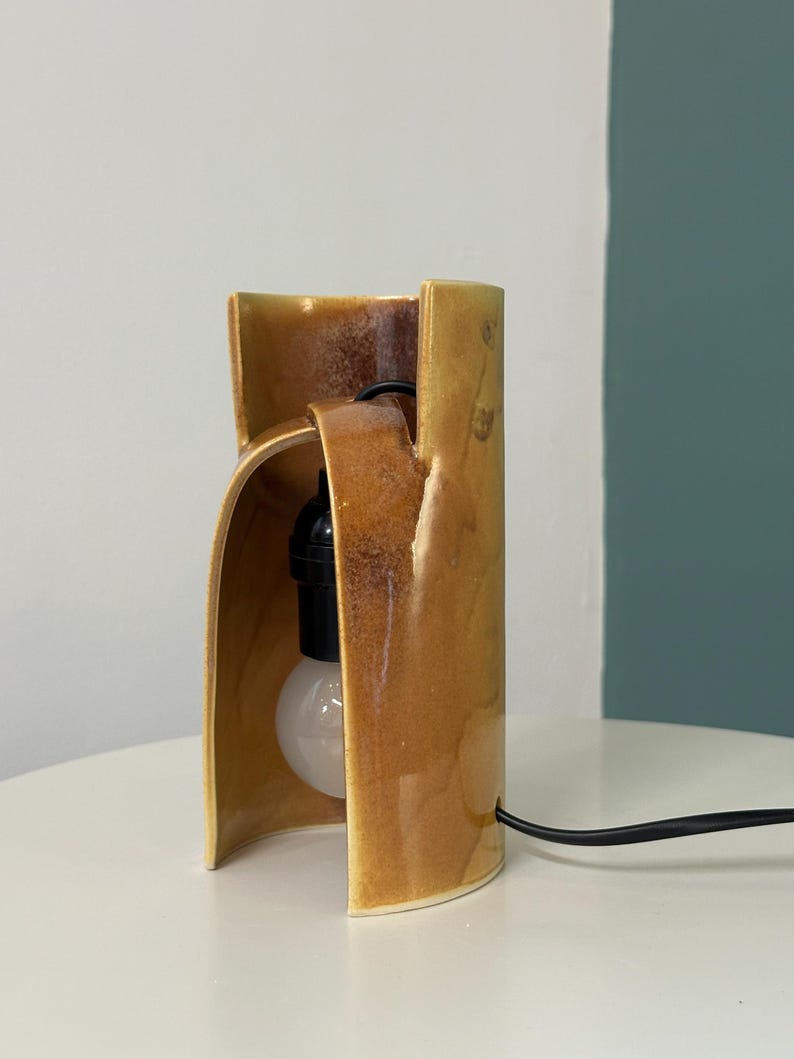 Puede incluir: Una l&aacute;mpara de mesa cil&iacute;ndrica de cer&aacute;mica con un esmalte amarillo mostaza brillante. La estructura presenta una abertura vertical que revela un casquillo negro y una bombilla redonda blanca. Un cable de alimentaci&oacute;n negro sale de la base.