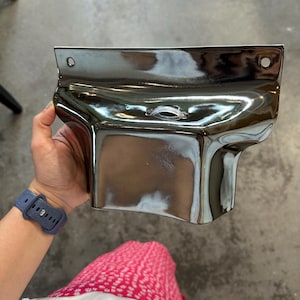 May include: A metallic, chrome-finished wall-mounted object with a curved, rectangular shape. It features a flat top edge with two circular mounting holes on the sides and a central horizontal opening with a clear plastic insert for cable management.