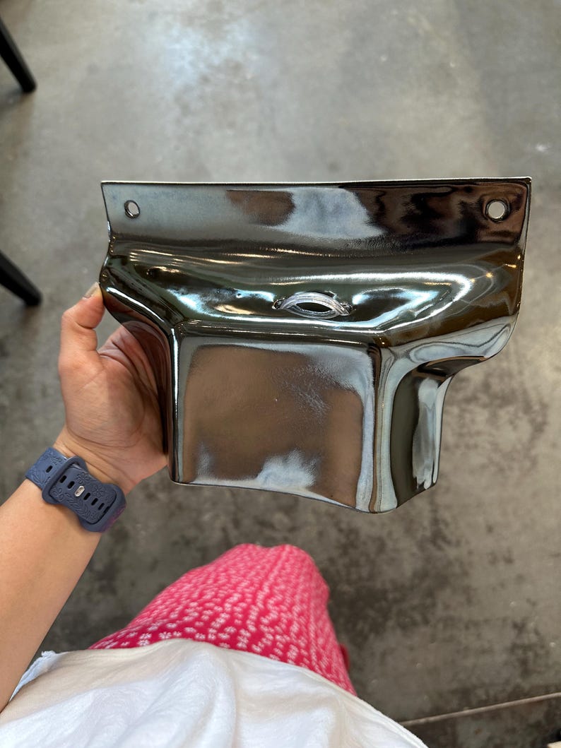 May include: A metallic, chrome-finished wall-mounted object with a curved, rectangular shape. It features a flat top edge with two circular mounting holes on the sides and a central horizontal opening with a clear plastic insert for cable management.