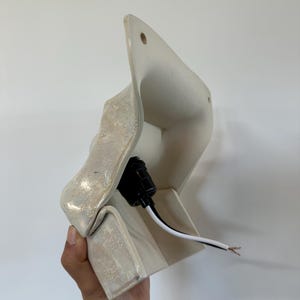 May include: A wall-mounted ceramic light fixture with an off-white, textured, iridescent glaze. The piece features a flared, angular shape with two small circular holes at the top for mounting and a black electrical socket with exposed wires.