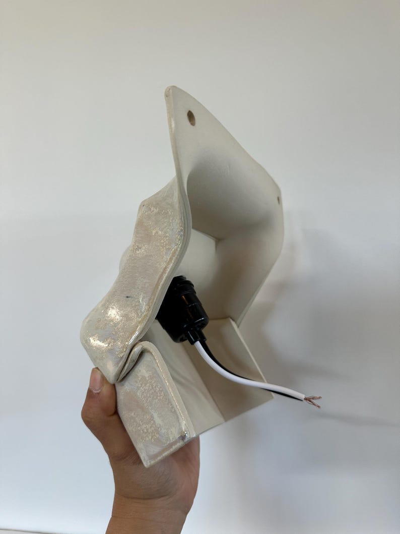 May include: A wall-mounted ceramic light fixture with an off-white, textured, iridescent glaze. The piece features a flared, angular shape with two small circular holes at the top for mounting and a black electrical socket with exposed wires.