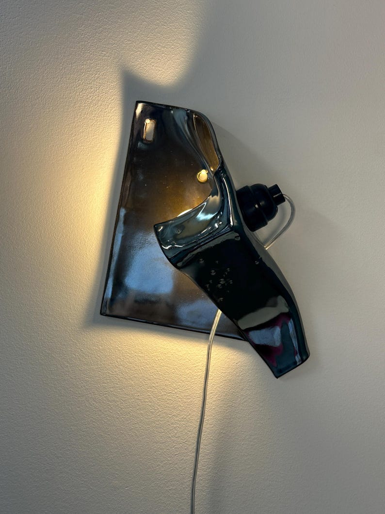 Peut inclure: Une applique murale compos&eacute;e d'un abat-jour en c&eacute;ramique gris anthracite brillant &agrave; la forme irr&eacute;guli&egrave;re et repli&eacute;e. Le luminaire comprend une douille noire et un cordon d'alimentation transparent, diffusant une lueur chaude et douce.