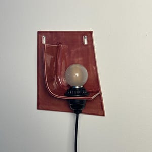 May include: A wall-mounted sconce featuring a glossy, terracotta-colored ceramic backplate with a curved, shelf-like projection. A black socket holds a round, frosted light bulb, and a black power cord extends downward from the base.