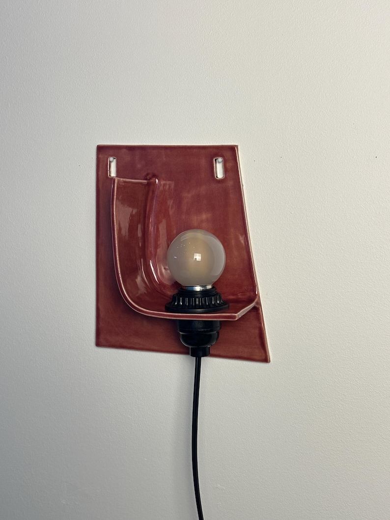 May include: A wall-mounted sconce featuring a glossy, terracotta-colored ceramic backplate with a curved, shelf-like projection. A black socket holds a round, frosted light bulb, and a black power cord extends downward from the base.