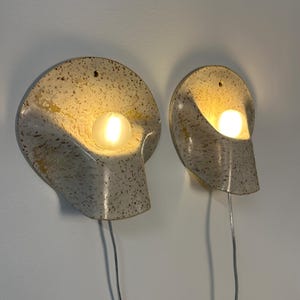 May include: Two wall-mounted sconces featuring a speckled cream and brown ceramic shell-like design. Each fixture holds a single glowing round light bulb and includes a small hole at the top for mounting, with a thin power cord extending downwards.