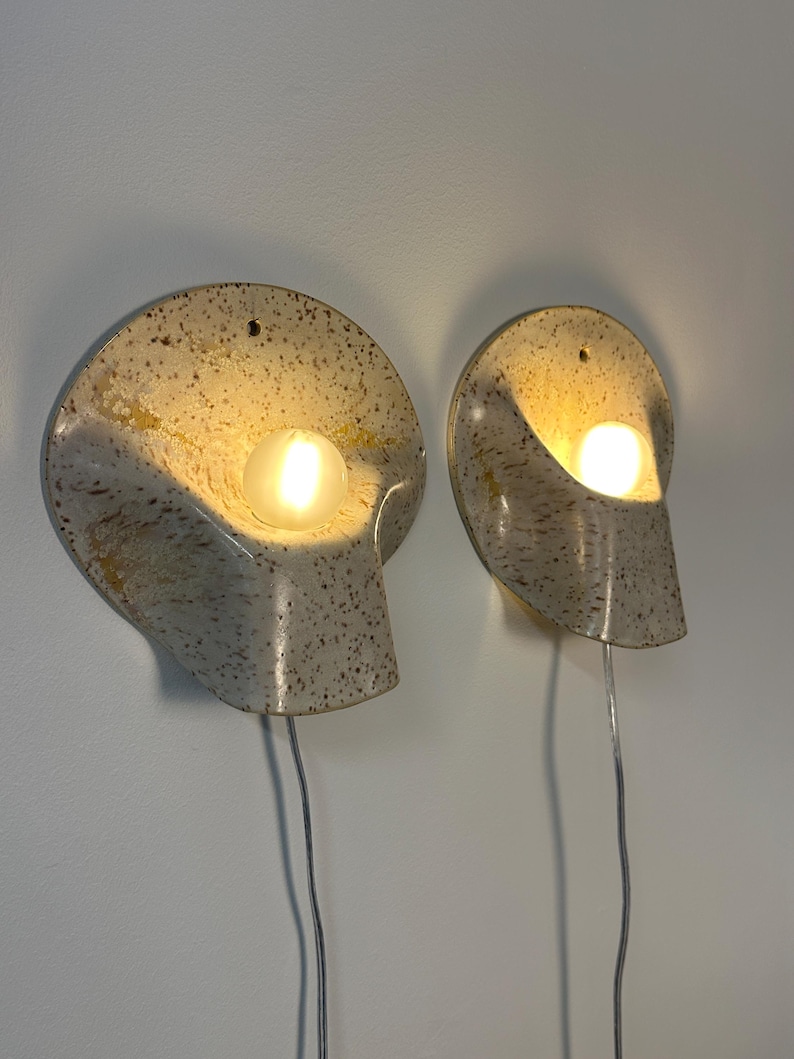 May include: Two wall-mounted sconces featuring a speckled cream and brown ceramic shell-like design. Each fixture holds a single glowing round light bulb and includes a small hole at the top for mounting, with a thin power cord extending downwards.