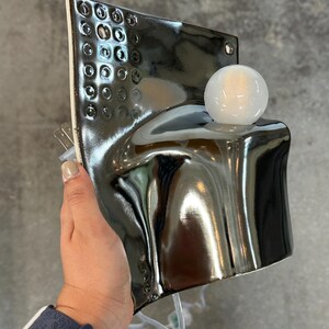K&ouml;nnte beinhalten: Eine Wandleuchte aus Keramik mit gl&auml;nzendem, dunkelmetallischem Finish und einem strukturierten Gittermuster im oberen Bereich. Die Lampe hat eine geschwungene, skulpturale Form und ist mit einer runden, wei&szlig;en Gl&uuml;hbirne aus Glas versehen.